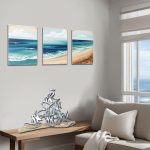 A modern coastal living room features a unique driftwood and metal sculpture of abstract fish on a wooden table, embodying driftwood art. Above the sculpture, three ocean-themed paintings with blue waves and sandy shores hang on the wall. The room has light gray walls, a large window with white shades allowing natural light and an ocean view, and a neutral sofa with soft pillows and a throw, creating a calm, beach-inspired atmosphere perfect for bathroom or living room decor.