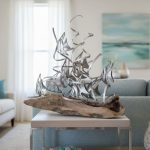 A modern driftwood and metal sculpture featuring abstract fish shapes sits atop a natural piece of driftwood on a light wooden table. The artwork is placed in a coastal living room with a soft blue sofa, white curtains letting in sunlight, and an ocean-inspired painting on the wall. The sculpture’s flowing metal lines and organic wood base create a nautical, beach-themed centerpiece that adds elegance and artistic flair to the serene, contemporary decor.
