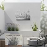 This image showcases a modern outdoor garden space featuring a sleek, minimalist gray wall with a decorative metal flower and grass wall art piece mounted in the center. The wall art is crafted from stainless steel, designed in a floral and grass motif, adding an artistic touch to the serene setting. Below the wall art, there is a built-in planter box filled with lush green grasses and tall, variegated plants, enhancing the natural ambiance. To the left, a comfortable outdoor sofa with gray cushions is placed, inviting relaxation. On the right, a black metal chair and a round, white fire pit filled with smooth stones create a cozy seating area. The background shows lush greenery and bamboo plants, further emphasizing the outdoor garden theme. Inset shows a close-up of the custom-sized small metal flower grass wall art, highlighting the intricate details of the floral design made from stainless steel, perfect for enhancing outdoor or garden decor with a modern, artistic touch. The overall scene embodies a stylish, tranquil outdoor environment with durable, artistic metal wall decor designed for outdoor use.