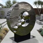 A sleek, contemporary outdoor metal sculpture featuring a large circular ring with a rippled, polished surface. Inside the ring are five shiny, reflective spheres arranged in decreasing size, creating a dynamic visual effect. The sculpture is made of high-quality, weather-resistant materials with a smooth, metallic finish. It is displayed on a black base in a modern outdoor setting, adding an artistic focal point to the landscaped environment.