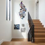 A modern interior staircase area with white walls and light wooden steps. The wall features a colorful, abstract metal wall art with swirling circles and layered shapes, creating a vibrant focal point. Adjacent to the staircase are three smaller framed artworks in various geometric and textured designs, adding an artistic and contemporary touch to the space. Natural light streams in through a tall window, brightening the clean and stylish decor.