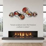 Decorative metal sculpture with circular and spherical elements mounted above a modern fireplace with a burning fire, on a white wall with the caption: 'Great sculpture over the fireplace crafted in metal.