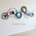 A modern, abstract wall art installation composed of multiple interconnected circular frames in various sizes and colors, with some containing colorful patterned panels and others featuring metallic spheres, mounted on a plain white wall above a light-colored wooden floor. The artwork is labeled "Custom Size" in the center.