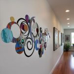 A contemporary interior hallway featuring a large, colorful, abstract metal wall art piece with circles, spirals, and layered shapes. The artwork adds a vibrant, artistic touch to a white wall. The hallway has wooden flooring, framed art on the wall, and a potted plant near a window, creating a bright, stylish, and inviting space.