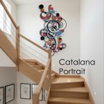 Modern abstract metal wall art sculpture titled “Catalana Portrait” displayed on a white wall above a wooden staircase. The art piece features colorful blue, orange, black, and silver geometric shapes, circles, and swirling accents, creating a vibrant and contemporary focal point. This art for staircase area adds a bold statement and enhances the modern interior decor, complementing the framed abstract artwork on the lower wall. Ideal for homes seeking unique and stylish staircase wall art.