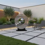 A modern outdoor garden space featuring a sleek, circular metal sculpture with a reflective, wavy surface and five polished spheres arranged along its inner edge. The sculpture is set on a black base and positioned on a paved patio with large square tiles. Surrounding the area are lush green plants, small trees, and a minimalistic lounge chair with a side table holding a potted plant, creating an elegant and contemporary outdoor setting.