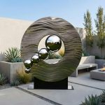 This modern outdoor sculpture features a circular, wave-like metal frame with a smooth, textured surface. Inside the circle are reflective stainless steel spheres of varying sizes, creating a striking visual contrast. Made from weather-resistant materials, it blends contemporary design with durability. Positioned in a minimalist garden setting, it serves as an eye-catching focal point that adds artistic sophistication and a sense of movement to outdoor spaces.