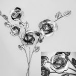 A handcrafted metal rose wall sculpture featuring intricately layered petals and detailed leaves in a polished silver finish. The reflective surfaces enhance depth and elegance, creating a modern floral art piece.