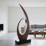 Modern abstract metal sculpture with curved design, displayed in a minimalist living room near gray sofa.