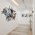 A hallway with light wood flooring displays a large, Picasso-inspired abstract metal wall sculpture featuring bold, fragmented faces and vibrant colors. Across from it, three framed prints in a Cubist style hang above a slim console table with a minimalist vase. Soft lighting and a neutral rug complete the modern, artistic look.
