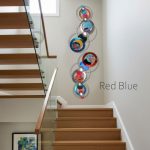A contemporary interior staircase features a modern wall art piece titled "Red Blue," composed of interconnected circular elements in shades of red, blue, and metallic, mounted vertically on the wall beside the staircase. The artwork includes abstract patterns within the circles and is complemented by the wooden staircase and glass railing in the space.