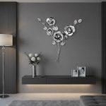 A modern, minimalist interior features a sleek, floating black shelf mounted on a gray wall. On the shelf, there is a tall silver vase with white roses and greenery, alongside two small framed black-and-white photographs. Above the shelf, a metallic wall decoration of three large, stylized flowers with leaves adds a decorative touch. To the left, a tall, white floor lamp with a slender stand casts a soft light. The room has a light-colored floor with a textured gray rug, and a portion of a gray upholstered furniture piece is visible in the bottom right corner. The overall aesthetic is elegant and contemporary, with a monochromatic color scheme accented by metallic and floral elements.