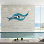 A contemporary indoor pool area with floor-to-ceiling windows overlooks a beach and palm trees. A striking abstract wall sculpture, featuring intertwined blue and silver metallic wave shapes with a circular mirror accent, is mounted on a large beige stone tile wall. Soft natural light fills the space, reflecting off the pool water. Minimalist decor includes a white sofa, a glass coffee table, and small green potted plants, creating a serene, coastal-inspired atmosphere.