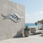 A contemporary outdoor patio area features a striking abstract metallic eye sculpture mounted on a tall concrete wall. The sculpture’s reflective surface and flowing design draw attention, while a large potted agave plant adds greenery in the foreground. To the right, a modern gray outdoor sofa with plush cushions invites relaxation. The background reveals a serene ocean view, blue sky, and palm trees, creating a peaceful and stylish seaside atmosphere.