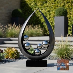 This modern sculpture features a sleek, black teardrop-shaped frame with polished, reflective metal spheres arranged inside. The artwork is set outdoors in a landscaped garden with lush green plants, ornamental grasses, and stepping stones, creating a contemporary aesthetic. The reflective surfaces of the spheres mirror the surrounding environment, including the sky, trees, and nearby structures, adding depth and visual interest to the installation. The sculpture stands on a sturdy black base, blending artistic elegance with outdoor durability.