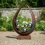 A modern outdoor sculpture features a rusted, teardrop-shaped metal frame with a weathered appearance, standing on a rectangular base. Inside the frame, shiny silver spheres of varying sizes are arranged in a balanced, artistic composition. The sculpture is set on a gravel surface, with a lush garden of green foliage, white flowers, and a stone wall in the background under a bright, clear sky. The reflective spheres mirror the surrounding environment, adding depth and visual interest.